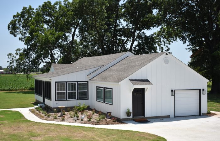 A white single-story house with a gray roof sits on a grassy lawn. There are large trees in the background and a small garden near the entrance.
