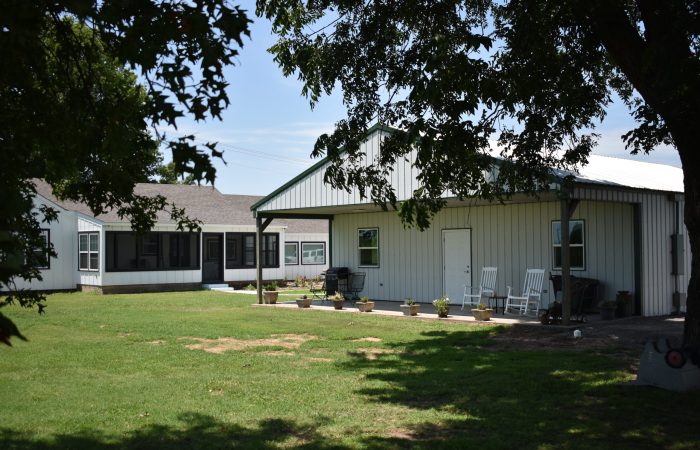 A white metal building and a gray-roofed house are surrounded by grass and trees. Two white chairs and several potted plants line the porch of the metal building.