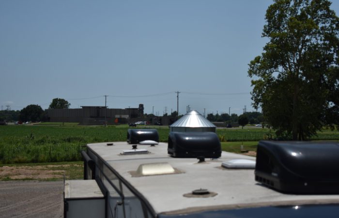 Rooftop view of a vehicle featuring ventilators, with a green field, a silo, and a distant building under a clear blue sky.