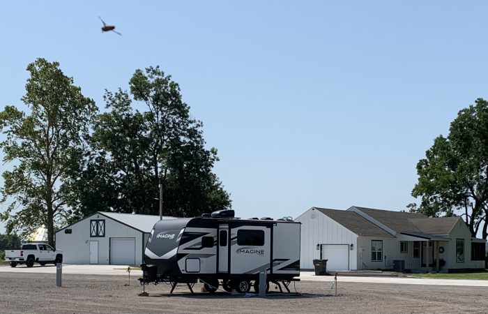 A parked RV trailer on gravel near a white house and barn, with a helicopter flying in the sky above. Trees and a parked truck are visible in the background.